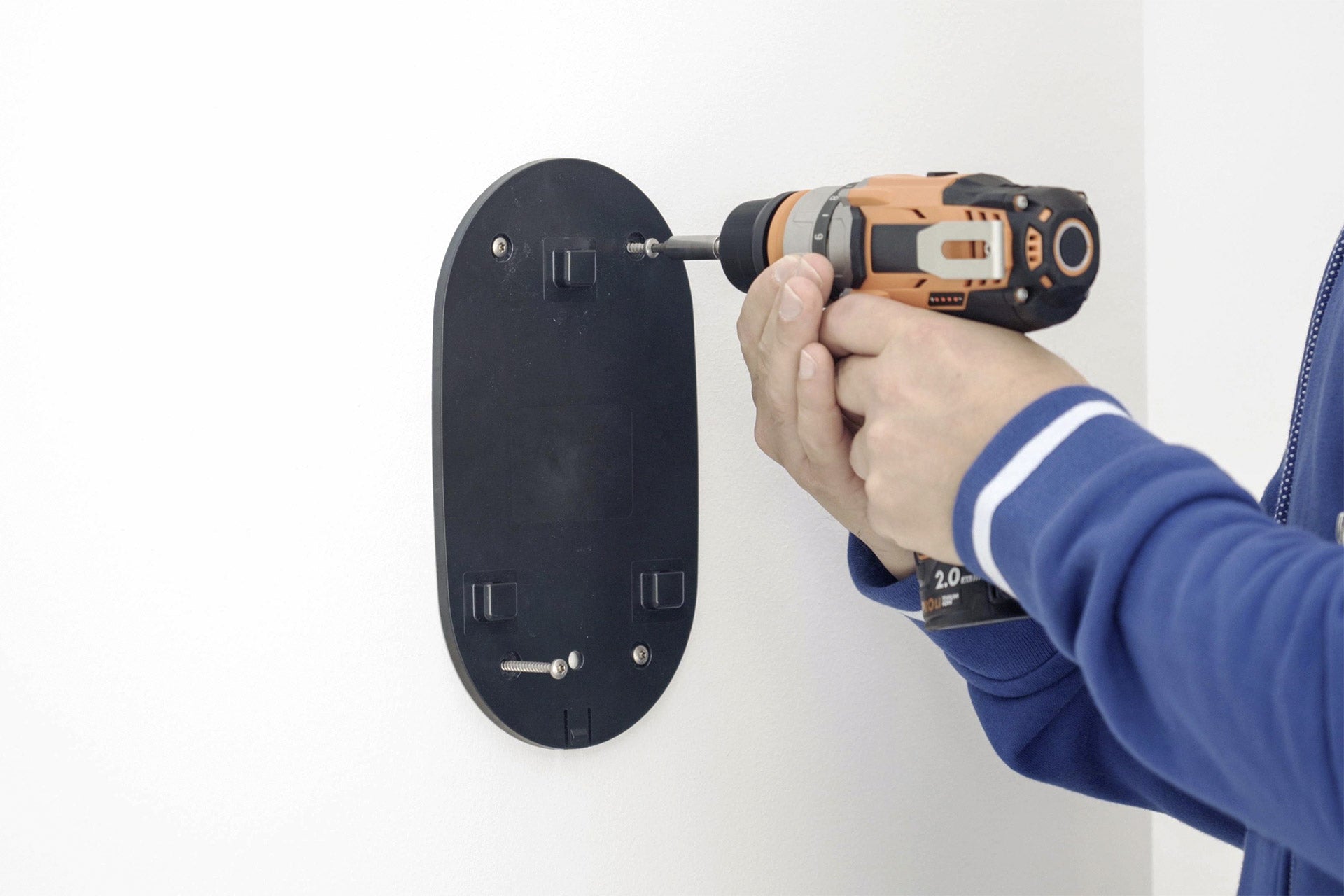 Eine Person befestigt mit einem Akkuschrauber eine schwarze go-e Charger Ersatz-Wandmontageplatte von go-e an einer weißen Wand.