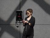 Eine Frau mit Sonnenbrille telefoniert, während sie neben einer GTÜ Wallbox WL60E-Ladestation für Elektrofahrzeuge steht und die auf ihrem Gerät angezeigte Ladeübersicht überprüft.